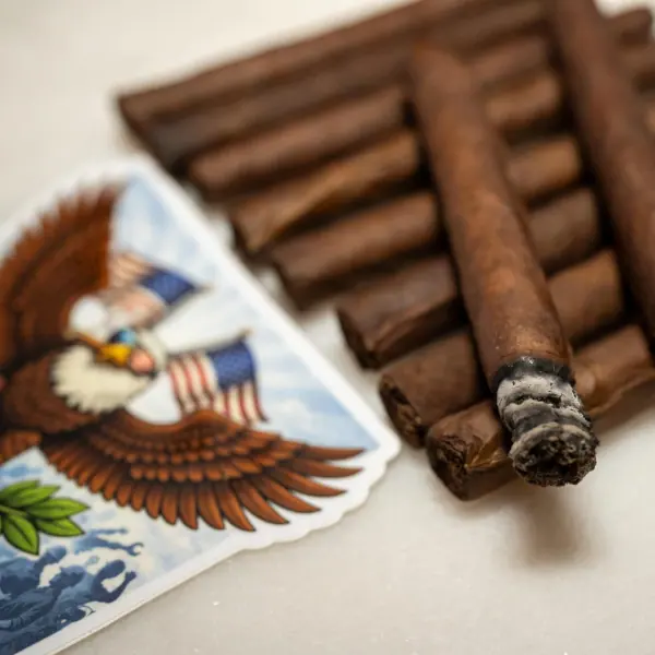 A freshly lit Blue Collar Tobacco cigar resting on a stack of hand-rolled cigars beside the eagle logo card